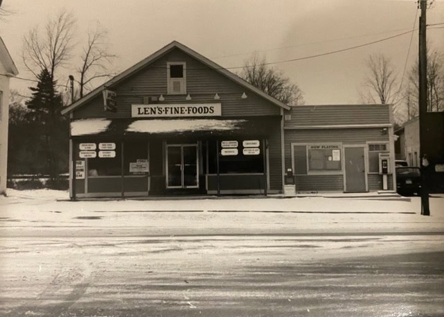 black and white photo of len's fine foods grocery store on a snowy day, with various signs on the storefront and bare trees in the background.