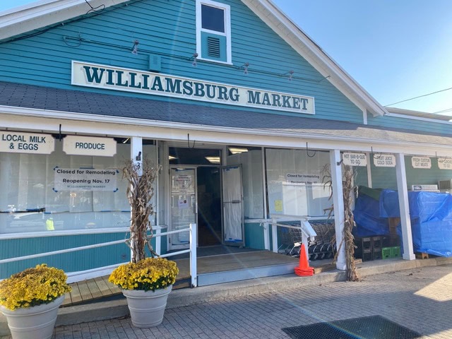 williamsburg market storefront with "closed for remodeling" signs, autumn decorations, yellow flowers, and blue tarps outside under a clear sky.