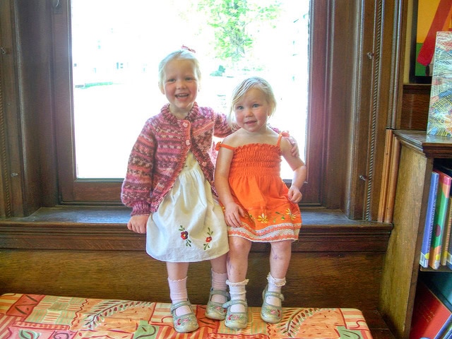 two young children smile while sitting on a wooden ledge by a large window. one wears a white dress with a pink sweater; the other wears an orange dress. books are visible on a nearby shelf.