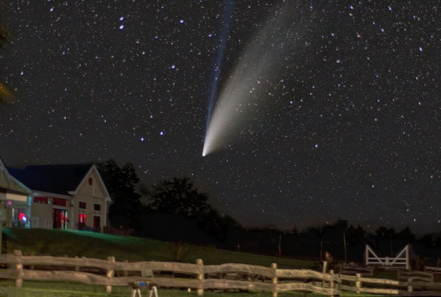 a bright comet with a long tail is visible in a star filled night sky above a rural landscape with a house, trees, and a wooden fence in the foreground.