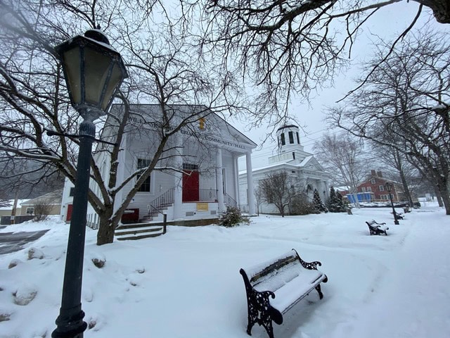 snow covered benches and trees line a quiet street with two white churches and a lamppost on a winter day.