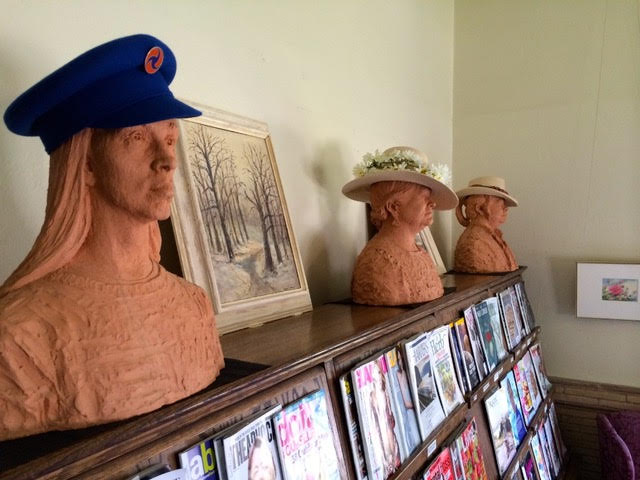 three clay busts wearing different hats are displayed on a wooden shelf above neatly arranged magazines and a framed landscape painting.