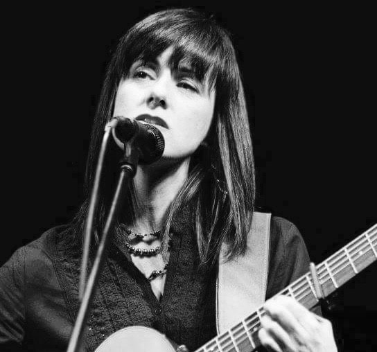 a woman with straight, shoulder length hair sings into a microphone while playing an acoustic guitar. the image is in black and white.