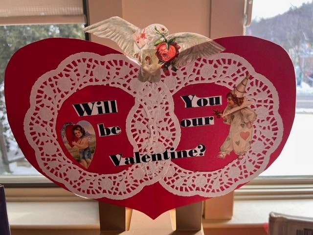 a red heart shaped valentine’s card with lace doilies, images of a dove, a boy, and a clown, and the text “will you be our valentine?” in black letters.