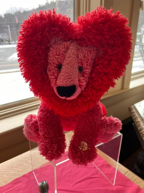 a plush toy shaped like a pink lion with a red, heart shaped mane sits on a clear display stand near a window.