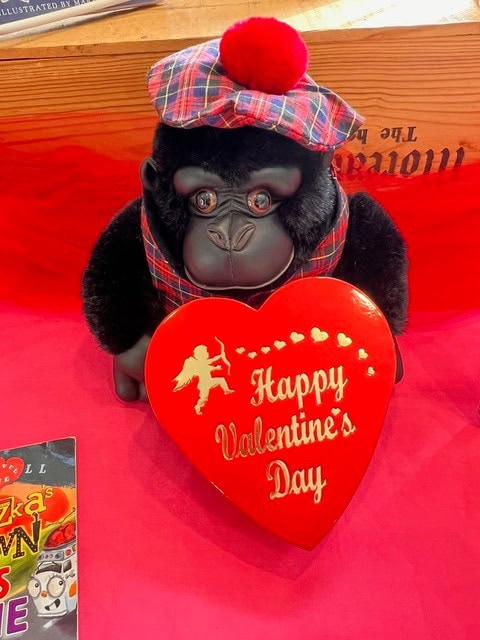 a plush gorilla wearing a plaid hat and vest holds a red heart shaped box that reads "happy valentine's day" in white lettering, with a pink background.