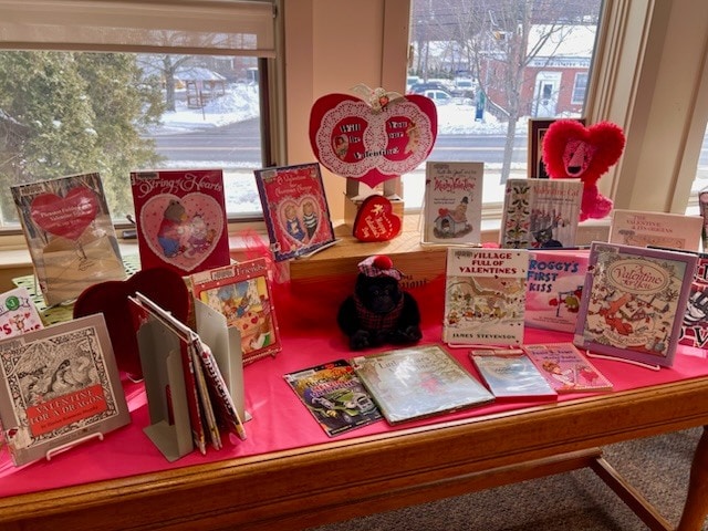 a display of valentine's day themed children's books and decorations is arranged on a table in front of a window with a snowy scene outside.