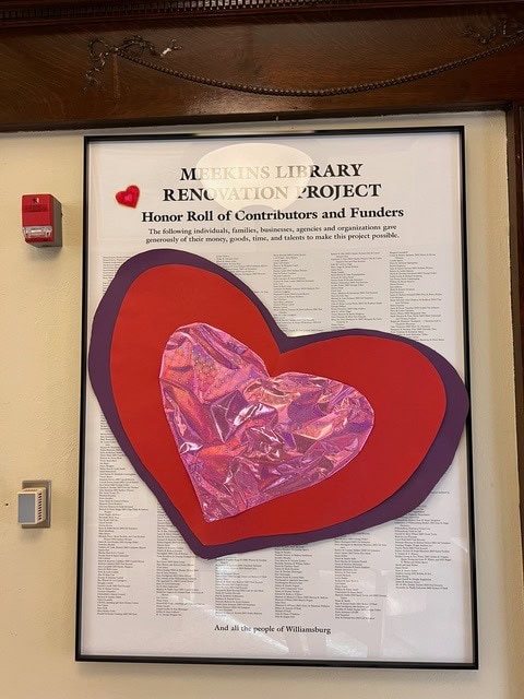 a framed poster titled "meekins library renovation project" lists contributors and funders, decorated with layered paper hearts in red and purple, and a small fire alarm nearby.