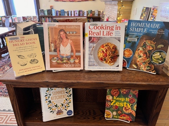 a display of seven cookbooks and food related books arranged on a wooden shelf in a library or bookstore setting.