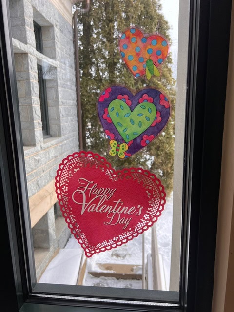 three colorful paper hearts, including a red one with "happy valentine's day" text, are taped to a window with a snowy outdoor view in the background.