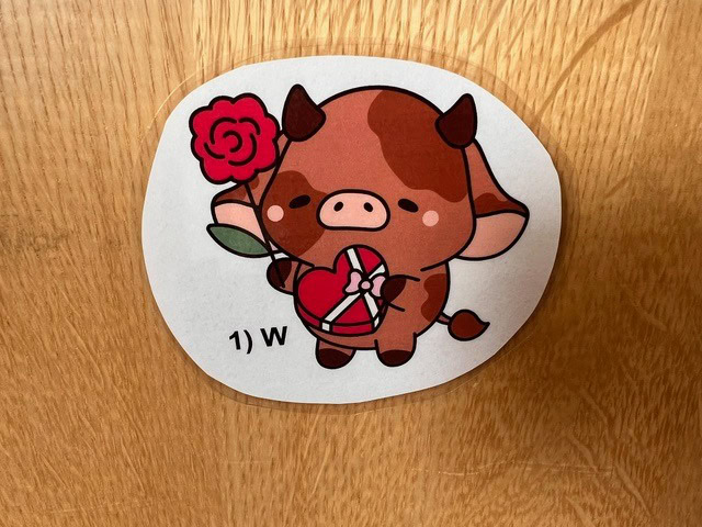 cartoon cow holding a red rose and a heart shaped box, with "1) w" written below, on a wooden surface.