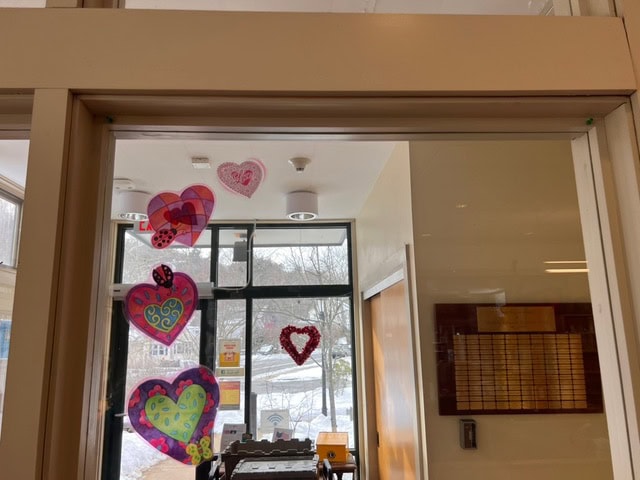 paper hearts are taped to a glass window above a doorway, with a large heart hanging on a window in the background. snow is visible outside.