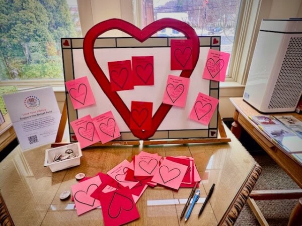 a display board with a large heart shape holds pink and red paper cards with heart outlines. more cards, pens, and small items are spread on the table in front of the display.