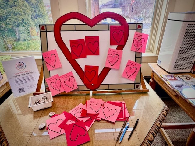 a display board with a large heart shape holds pink and red paper cards with heart outlines. more cards, pens, and small items are spread on the table in front of the display.