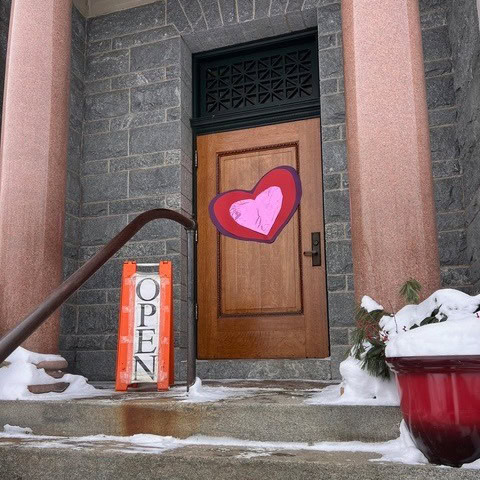 a wooden door with a large heart decoration is flanked by red columns, an "open" sign, and a snow covered red planter on stone steps.