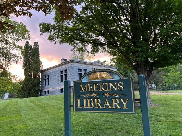 a green and gold sign reading "meekins library" stands on a grassy lawn in front of a stone building surrounded by trees at sunset.