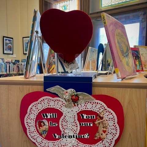 a library display features a large red heart, books, and a decorated sign that reads, "will you be our valentine?" with vintage valentine images.
