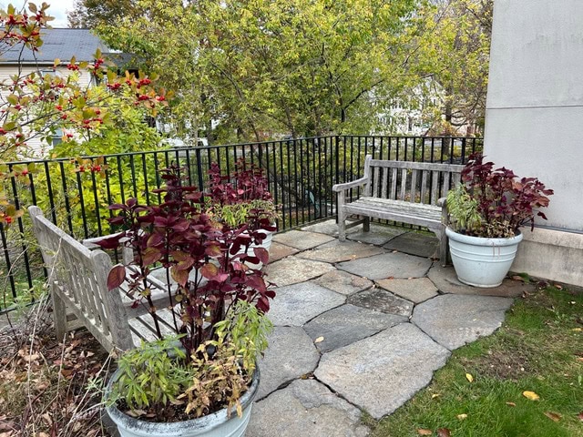 stone patio with two wooden benches and large planters containing red and green foliage, surrounded by a black metal railing and trees in the background.