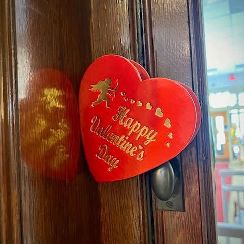 a red heart shaped sign with gold text reading "happy valentine's day" and a cupid graphic hangs on a wooden door.