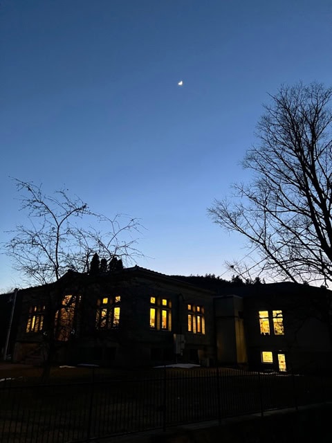 a building with illuminated windows stands under a twilight sky with a crescent moon visible; bare trees frame the scene.