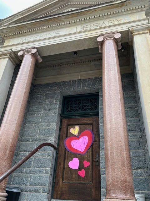 the entrance of meekins library, featuring stone columns and a wooden door decorated with colorful paper hearts.