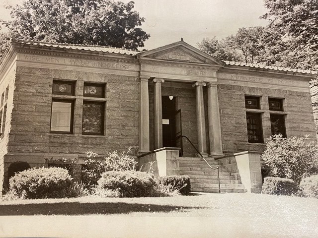 black and white photo of a small stone library building with columns, large windows, and a sign above the entrance. bushes and trees surround the front lawn.