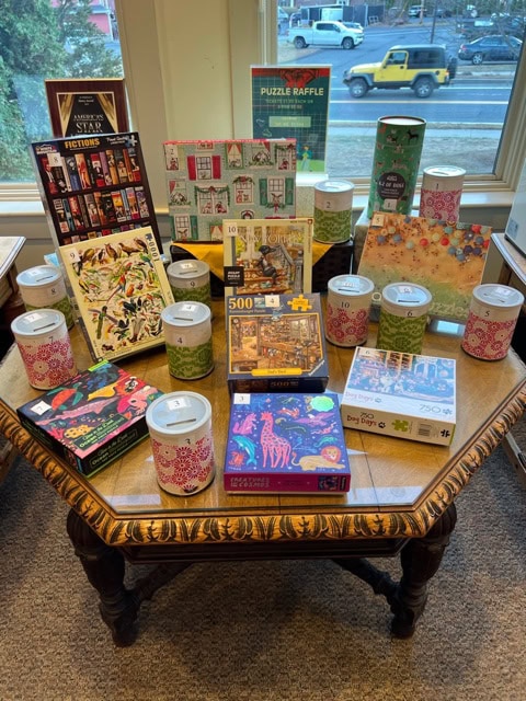 a wooden table displays various jigsaw puzzles and raffle jars with numbered labels, set in front of large windows showing a street outside.