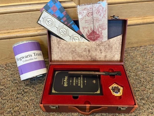 a harry potter themed collectible set including a trunk, journal, chocolate frog box, cards, and a canister labeled "hogwarts trunks collectible set," displayed on a carpeted floor.