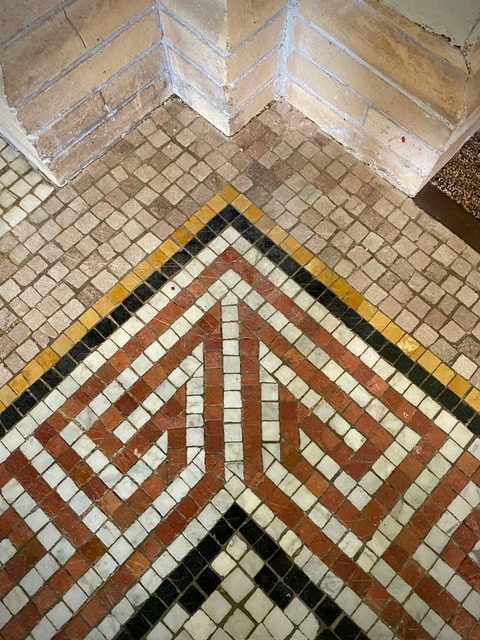 corner view of a tiled floor with a geometric mosaic pattern in red, black, white, and yellow, set against a tan stone wall.