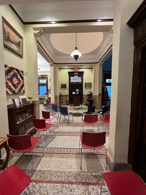 a room with red chairs arranged in rows facing a podium, featuring mosaic tile flooring, bookshelves, and framed artwork on the walls.