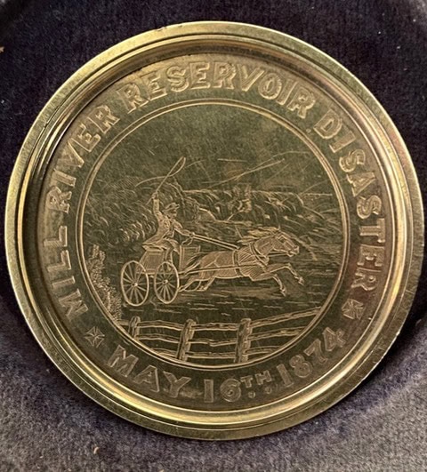 a commemorative medallion engraved with "mill river reservoir disaster may 16th, 1874" and an image of a person in a horse drawn carriage fleeing a flood.