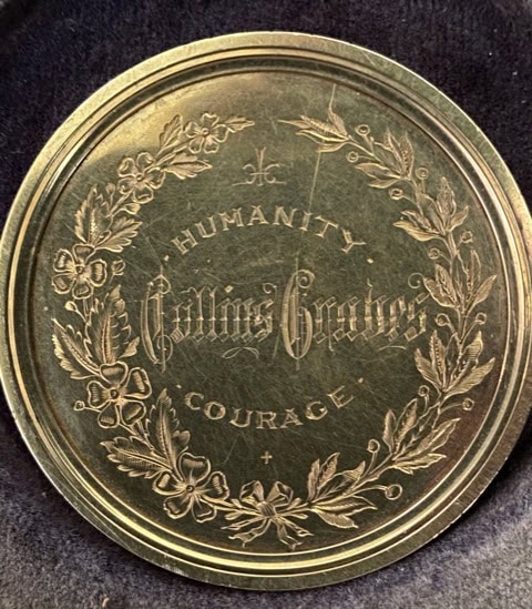 a round metallic medallion engraved with “humanity courage” and “collins graves,” surrounded by floral and leaf motifs.