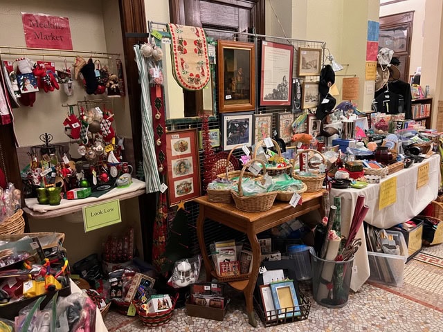 a craft sale stall displaying holiday ornaments, framed art, woven baskets, and various knick knacks on tables and wire grid walls.