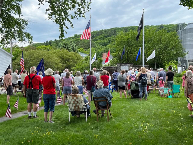a group of people stands and sits on chairs outdoors, facing several flagpoles and a memorial, with american flags visible around the area.