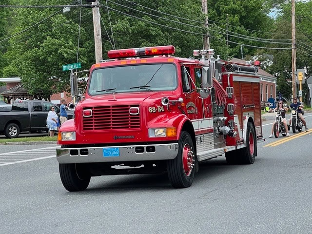 a red fire truck drives down a street during a parade, with people and motorcycles visible in the background.
