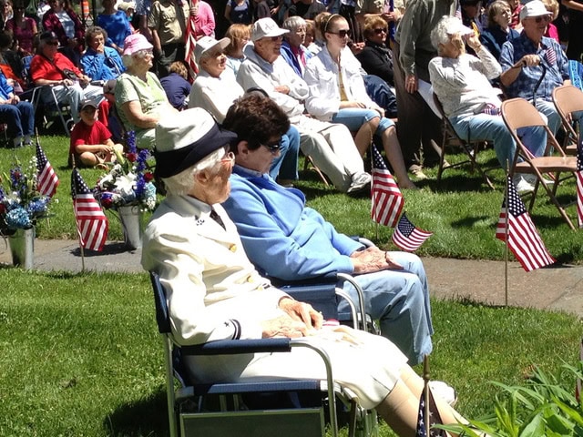 elderly people seated outdoors at an event, surrounded by american flags and flower arrangements, with a crowd watching in the background.