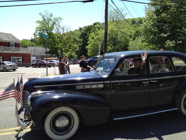 a vintage black car with american flags on the front is driving down a street during a parade; a person inside waves while onlookers stand nearby.
