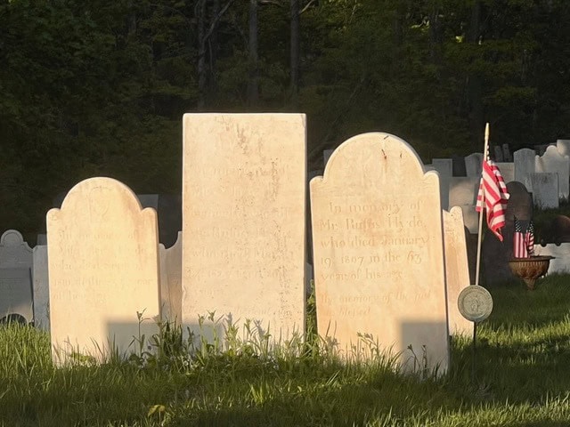 three old headstones stand in a sunlit cemetery, with an american flag and a circular medallion placed beside the rightmost marker.