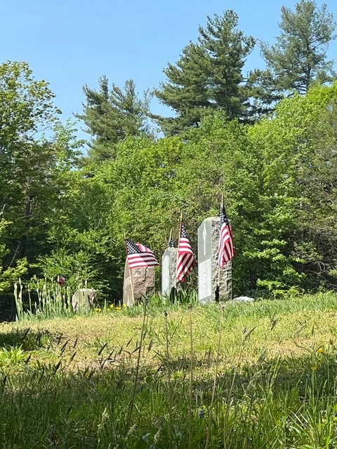 three gravestones with american flags stand in a grassy cemetery surrounded by green trees under a clear blue sky.