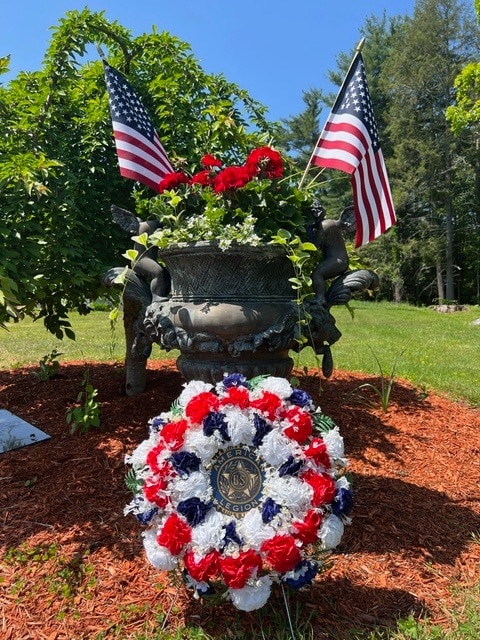 a patriotic flower arrangement with red, white, and blue flowers and two american flags is placed in front of a us legion emblem outdoors on a sunny day.