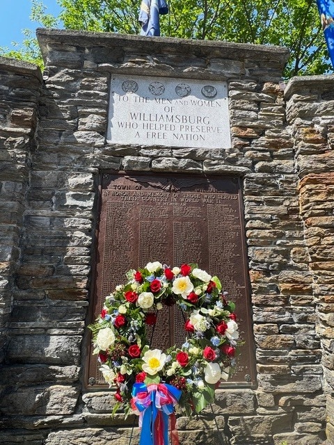 a stone memorial wall with plaques honoring williamsburg men and women who served, adorned with a wreath of red, white, and yellow flowers and blue and red ribbons.