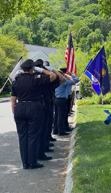 a group of uniformed officers stand in a line on a roadside, saluting while holding american and organizational flags on a sunny day.
