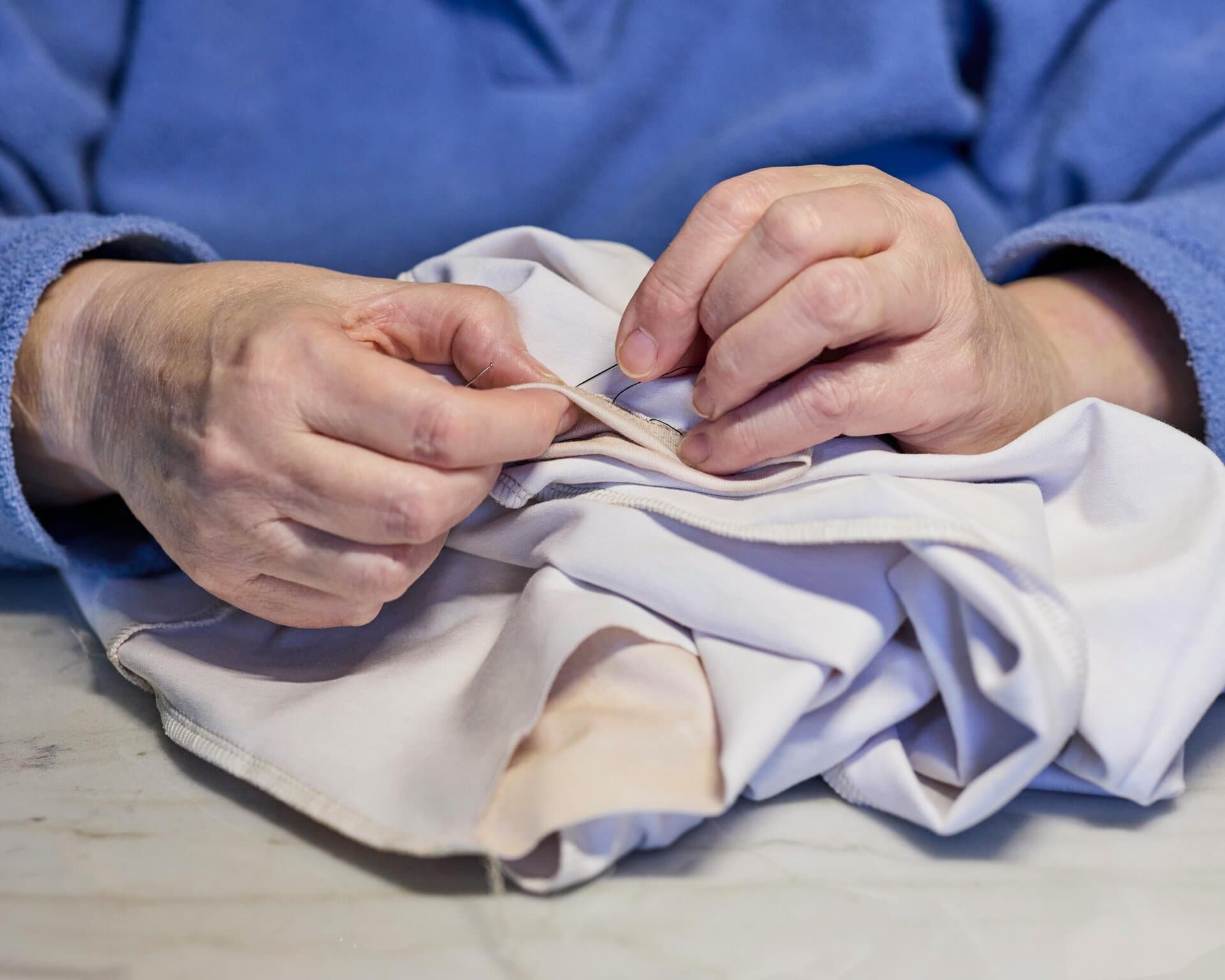 a person in a blue top is hand sewing a piece of light colored fabric using a needle and thread.