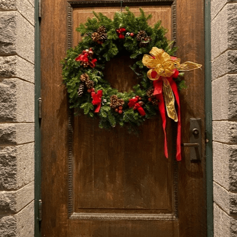a wooden door is decorated with a green holiday wreath adorned with pine cones, red berries, red ribbons, and a large gold and red bow on the right side.