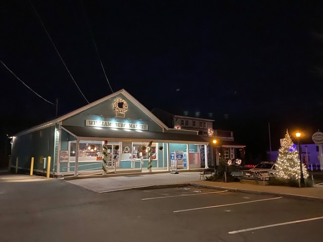 a small market building with a holiday wreath and lit christmas tree outside, pictured at night with few cars in the parking lot.