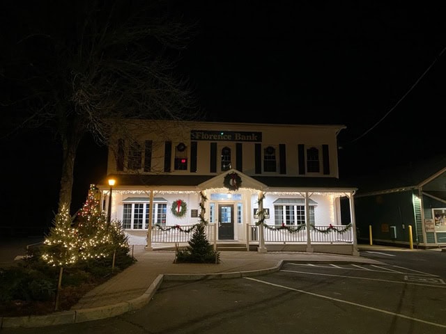 florence bank building at night, decorated with white holiday lights, wreaths, and small lit christmas trees in front.