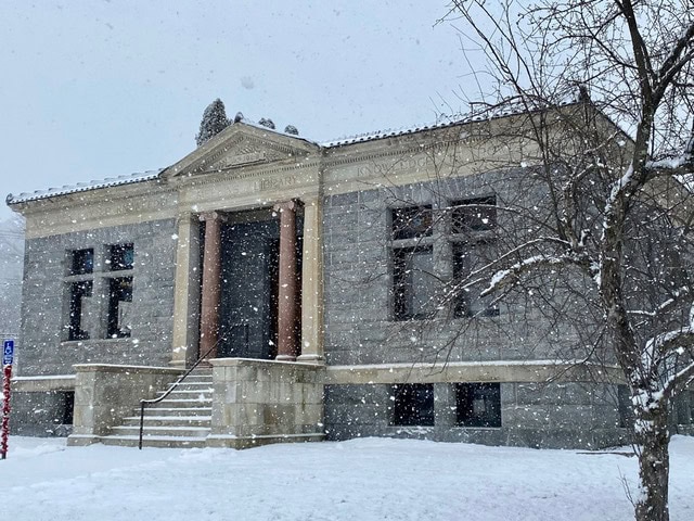 a stone building with columns at the entrance is seen during snowfall, with snow covering the ground, steps, and surrounding trees.
