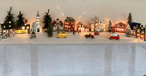a miniature christmas village display with lit buildings, trees, cars, and figurines arranged on a white cloth.