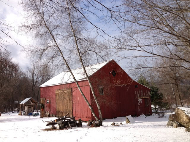a red wooden barn with weathered doors stands on a snowy landscape, surrounded by bare trees and a small wooden shed in the background.