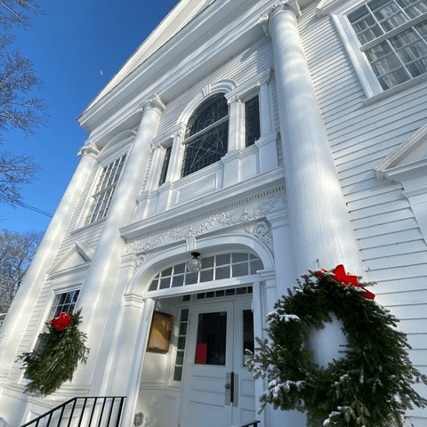 white colonial style building with large columns, adorned with two green wreaths with red bows, viewed from below on a clear winter day.
